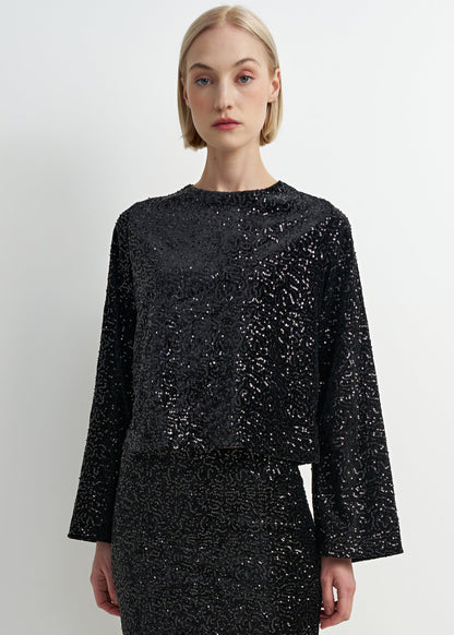 A model with short blonde hair wearing a black long-sleeve crop top and matching skirt set, both heavily embellished with silver sequins, against a plain light gray background.