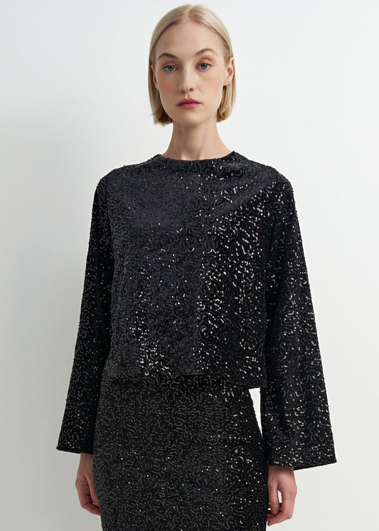 A model with short blonde hair wearing a black long-sleeve crop top and matching skirt set, both heavily embellished with silver sequins, against a plain light gray background.