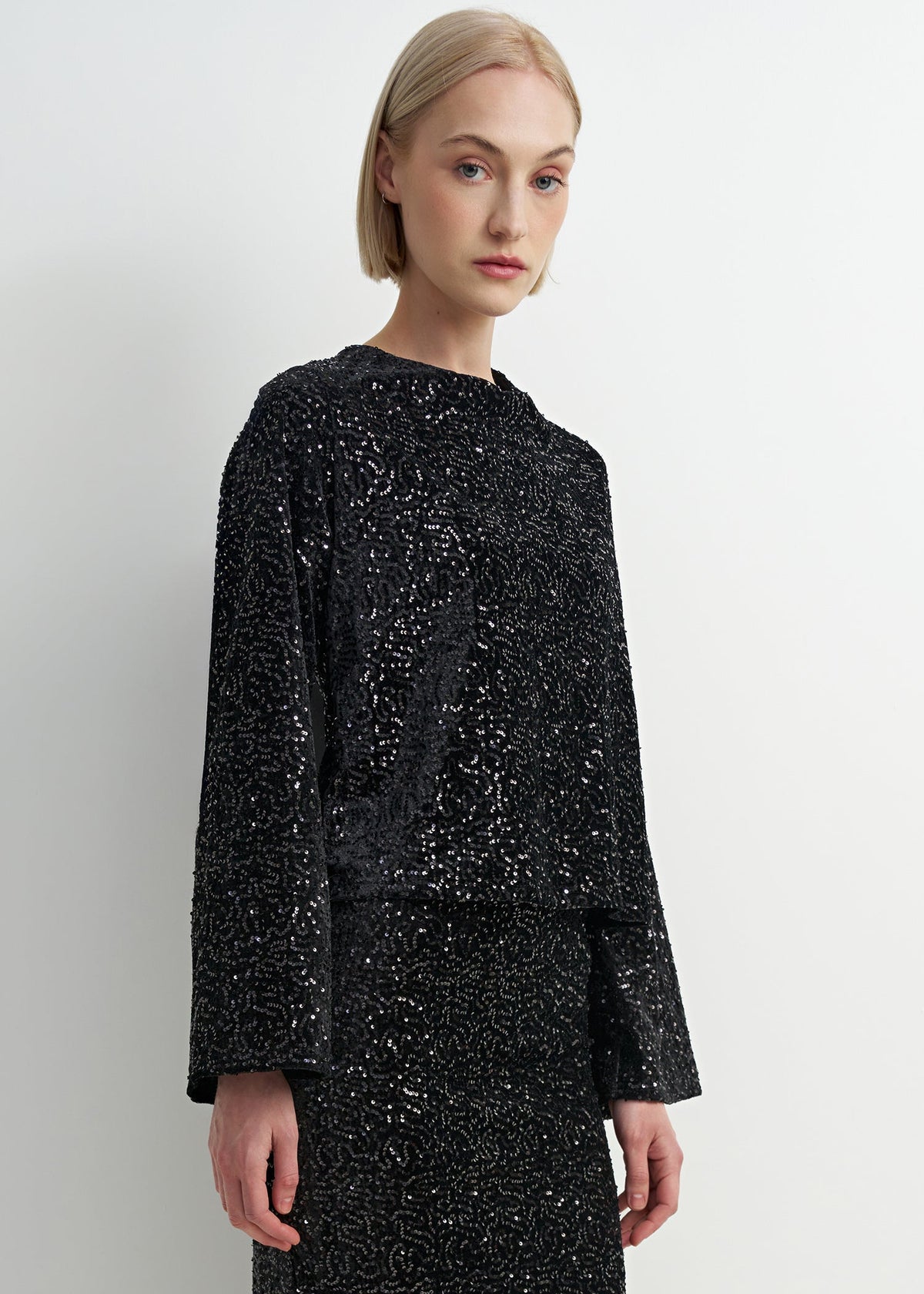 A model with short blonde hair wearing a black long-sleeve top and matching skirt, both heavily embellished with silver sequins, against a plain light gray background.