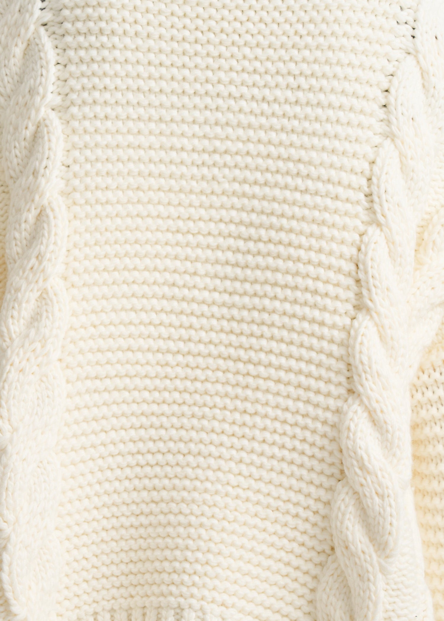 Close-up of a cream-colored cable-knit sweater showing the detailed knit texture and twisted cable patterns.