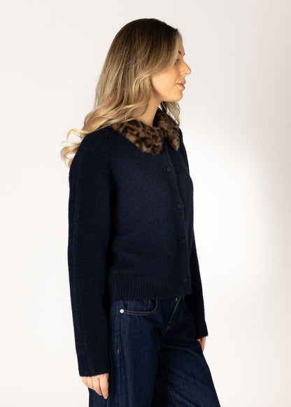 A woman shown in profile wearing a dark navy knit cardigan with a brown and black leopard-print collar, paired with dark blue jeans, against a plain off-white studio background.