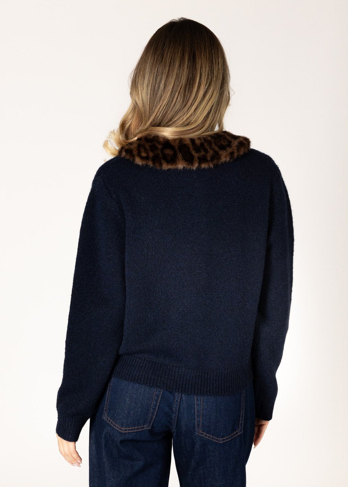 A woman viewed from behind wearing a navy blue knit sweater with a brown and black leopard-print faux fur collar, paired with dark blue jeans, against a plain off-white background.