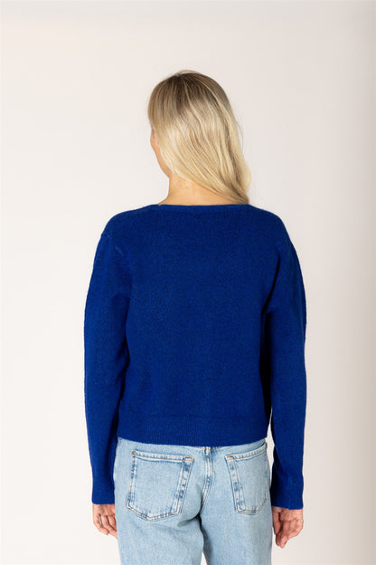 A woman viewed from the back wearing a solid royal blue knit sweater and light wash blue jeans, standing against a plain off-white studio background.