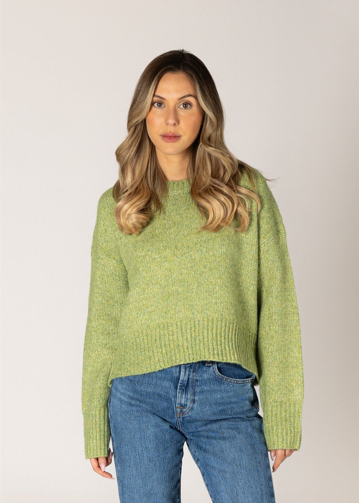 A woman with blonde-highlighted hair wearing a light green knit sweater and blue jeans, standing against a plain light gray studio background.