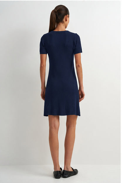 A woman viewed from the back wearing a short-sleeve ribbed knit dress in dark navy blue, paired with black flats, against a plain light gray studio background.